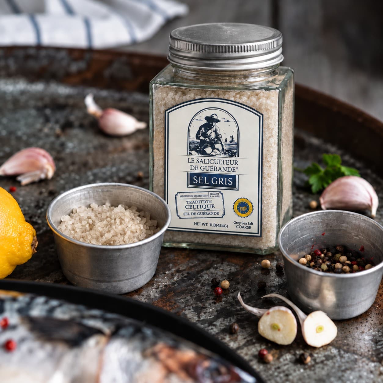 Le bocal de sel celtique gros avec de l’ail, du citron, des grains de poivre et du poisson sur un plateau rustique